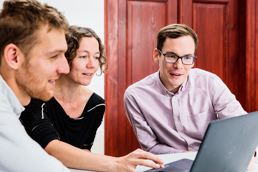 Drei Personen sitzen vor nebeneinander an einem Tisch und schauen auf einen Laptop